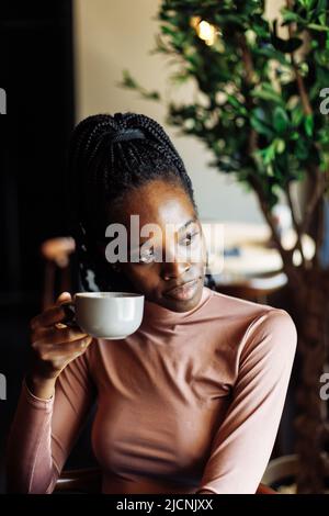 Vertical photo calm, dreaming african afro american young woman with dreadlocks, drinking coffee in cafe. Coffee break Stock Photo