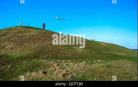 Wiltshire,England,UK-February 18th 2021: A little boy watches his Dad running along the top of the windswept Neolithic long barrow,dragging a kite hel Stock Photo