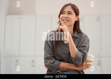 Portrait of a beautiful charming millennial Asian female freelancer or female entrepreneur with a coffee cup, hand on chin standing in the locker room Stock Photo