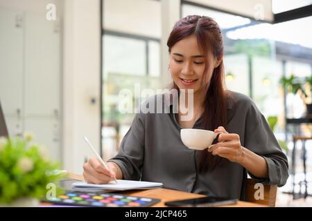 Relaxed young asian female graphic designer enjoy sipping coffee during coffee break and working in her office working desk. Stock Photo