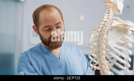 Male osteopath explaining bones on human skeleton exam, pointing at spinal cord to find anatomy diagnosis at physical therapy appointment. Therapist using spine to explain back pain. Close up. Stock Photo