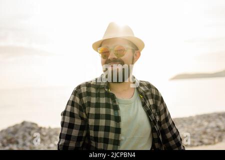 Handsome and confident. Outdoor portrait of smiling man wearing hat and sunglasses on beach. Holidays travel and summer tourism Stock Photo