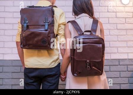 Back of woman and man with brown leather backpack on the brick wall background. Man and woman holding hands and presenting leather bags. Unisex bag fo Stock Photo
