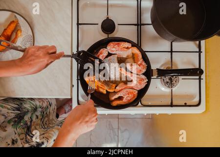 Top down view Hands with a fork and a skimmer frying salmon fish in a hot pan on a gas stove at home Stock Photo