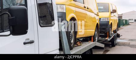 White small cargo truck car carrier loaded with two yellow van minibus on flatbed platform and semi trailer tow on roadside highway road. Volunteer Stock Photo