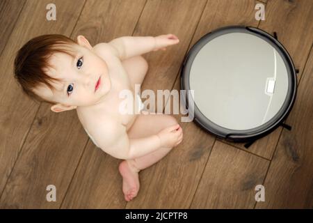 Happy toddler baby boy is playing with a robot vacuum cleaner on the floor. Kid aged one year Stock Photo