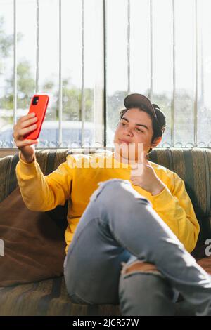 Friendly 20s young person with black cap on, wearing yellow color top t-shirt taking selfie on cell phone post. Photo on social network waving hand is Stock Photo