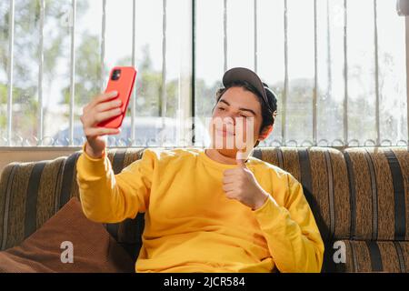 Friendly 20s young person with black cap on, wearing yellow color top t-shirt taking selfie on cell phone post. Photo on social network waving hand is Stock Photo
