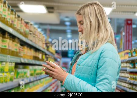 Young blonde woman reads the composition on a can of canned peas or corn in a store. Concept of buying food, proper nutrition Stock Photo