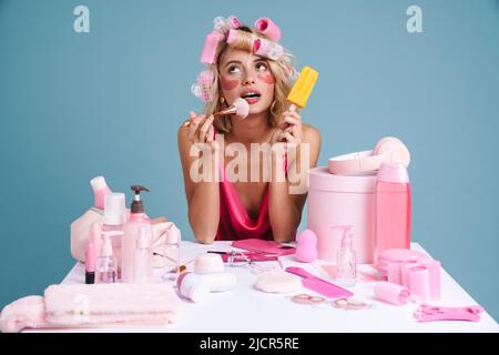 Young woman with curlers eating ice cream while doing makeup isolated over blue wall Stock Photo