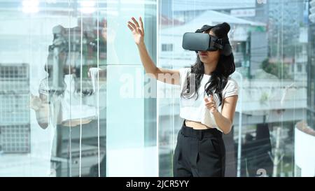 Excited businesswoman wearing virtual reality glasses, interacting with digital interface while standing near in large window at modern office. Stock Photo