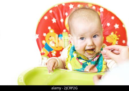 mother feeds funny baby from a spoon on a white background. child eats in a highchair Stock Photo