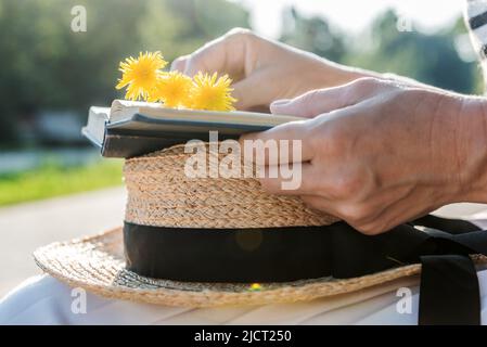 Hands of a woman holding a boater, a book and a bouquet of dandelions on her knees against blurred background of the park, close-up.Vacation concept. Stock Photo