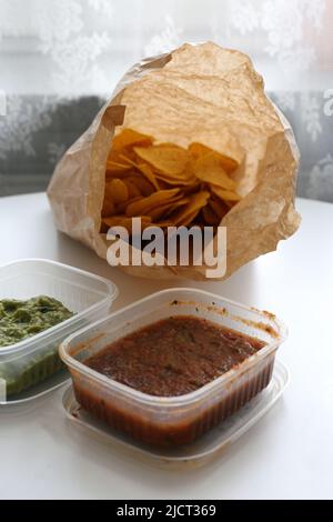 Mexican takeaway appetizer food on a table. There is a box of salsa, a box of guacamole and a bag of nachos to go taken to home. Delicious snack! Stock Photo