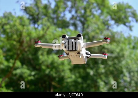 Small white mini drone flying up in front of forest, shallow depth of field Stock Photo