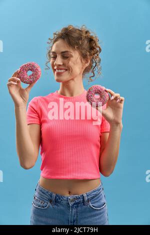 Cheerful woman with closed eyes, holding two colorful donuts, smiling in studio. Front view of curly-haired woman with pink sprinkled donuts, isolated on azure background. Concept of amusement. Stock Photo