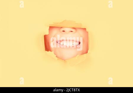 Positive child with toothy pleasant smile on face, keeps through torn hole in yellow paper. Breaking paper background. Emotions concept. Kid with Stock Photo