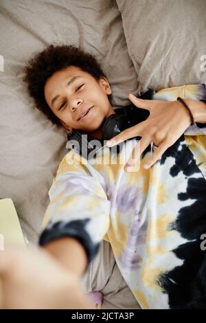 High angle POV shot of trendy black teenager wearing tie dye shirt while lying on bed and recording video for social media Stock Photo