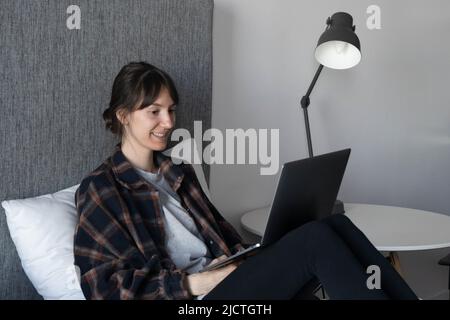 A young woman sits on the bed and smiling at home and pleasantly communicates with friends and relatives or spends time watching a movie. Lifestyle . Stock Photo