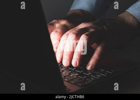 The hands of an unrecognizable Caucasian man in a blue shirt typing on a laptop keyboard. The scene is dark and the hands are illuminated by the light Stock Photo