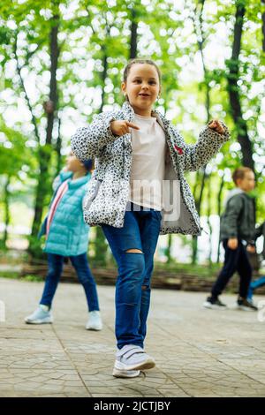 Cool little girl walking with other children in playground in green park. Summer holidays in camp, tourist center. Walking and playing outdoors, sport Stock Photo