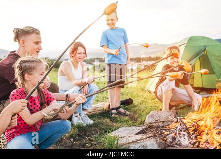 Group of six Kids: Boys and girls cheerfully smiling and roasting sausages on sticks over a campfire flame near the green tent. Outdoor active time sp Stock Photo