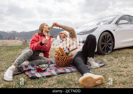 Young modern couple enjoying a picnic in nature, sitting near the parked white electric car Stock Photo