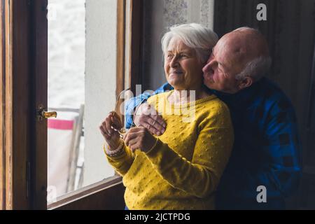 nice cheerful senior Caucasian couple standing near the window door and looking outside medium shoot indoor senior people love concept. High quality photo Stock Photo