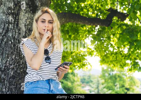 blond Caucasian girl holding a phone and showing silence gesture in the park. medium shot outdoor. High quality photo Stock Photo