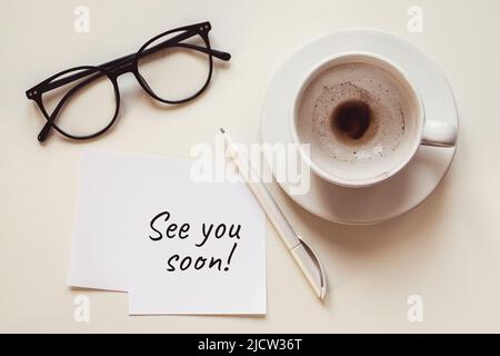 See you soon note, empty cup with coffee leftovers, eyeglasses and pen on white desk Stock Photo