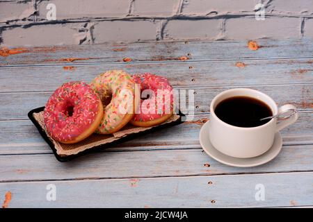 Three donuts in white and pink glaze with topping on a wooden table, next to a cup of black coffee. Close-up. Stock Photo
