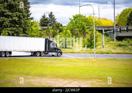 Loaded big rig black semi truck tractor transporting commercial cargo in dry van semi trailer running on the highway road at the city limit going on i Stock Photo