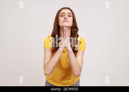 Please, I'm begging forgive. Adorable upset worried woman of young age with wavy hair looking with imploring desperate grimace, praying for help. Indoor studio shot isolated on gray background. Stock Photo