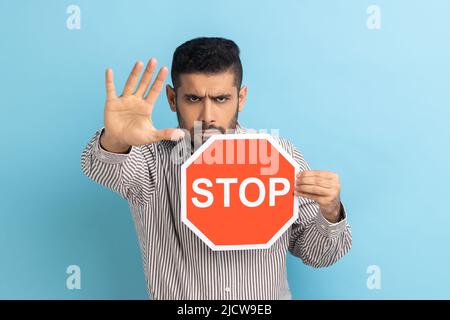 Businessman holding octagonal Stop symbol, showing red traffic sign and ban gesture with palm, warning of forbidden way, wearing striped shirt. Indoor studio shot isolated on blue background. Stock Photo