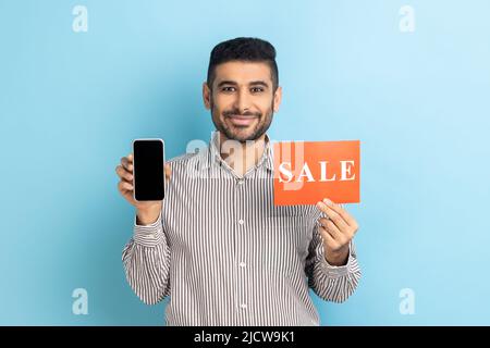 Cheerful businessman holding sale inscription, cellphone and smiling at camera, showing discounts on mobile device, application, wearing striped shirt. Indoor studio shot isolated on blue background. Stock Photo