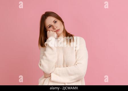 Portrait of blond woman leaning on hand and expressing disinterest apathy with dull look, feeling lazy indifferent, wearing white sweater. Indoor studio shot isolated on pink background. Stock Photo