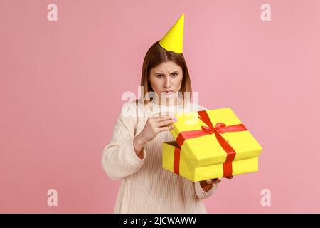 Portrait of sad upset unhappy blond woman in party cone unpacking present box, being disappointed with gift, looking at camera, wearing white sweater. Indoor studio shot isolated on pink background. Stock Photo