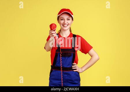 Portrait of affirmative worker woman holding out handset, asking call her and order service, looking friendly at camera, wearing overalls and red cap. Indoor studio shot isolated on yellow background. Stock Photo