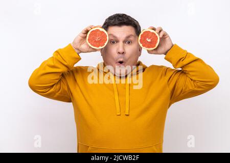 Portrait of excited middle aged man having fun, holding two halves of grapefruit, expressing astonishment, wearing urban style hoodie. Indoor studio shot isolated on white background. Stock Photo