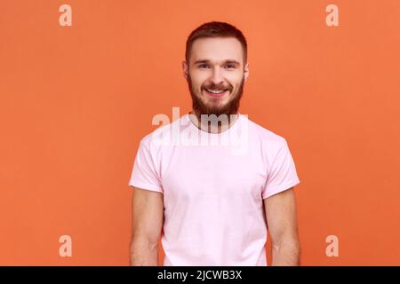 Portrait of happy bearded man looking at camera and smiling, expressing positive emotions, being in good mood, wearing pink T-shirt. Indoor studio shot isolated on orange background. Stock Photo