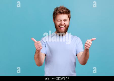 Portrait of bearded man approves incredible promo keeps thumbs up likes and agrees being satisfied with something, winking to camera. Indoor studio shot isolated on blue background. Stock Photo