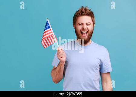 Portrait of overjoyed bearded man holding in hand flag of united states of america, celebrating independence day, having excited facial expression. Indoor studio shot isolated on blue background. Stock Photo