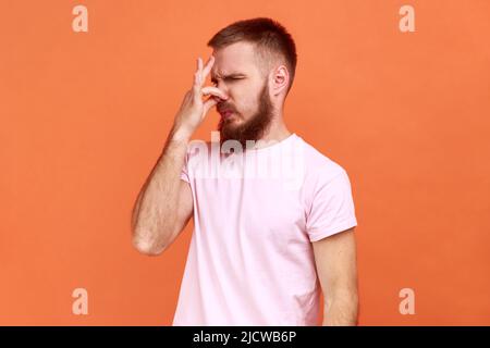 Portrait of confused bearded man grabbing nose, grimacing in disgust and expressing repulsion to stink, fart gases, wearing pink T-shirt. Indoor studio shot isolated on orange background. Stock Photo