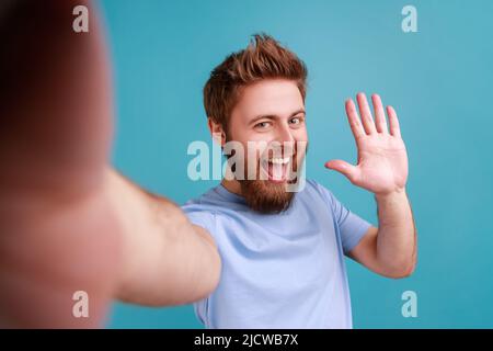 Portrait of positive friendly bearded man blogger making selfie or streaming, waving hand POV, point of view of photo, saying hello or bye. Indoor studio shot isolated on blue background. Stock Photo
