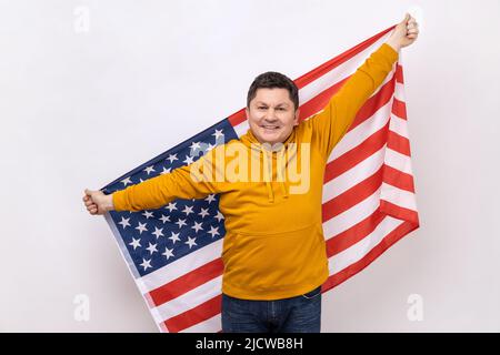 Happy positive middle aged man standing with raised arms, holding USA flag, celebrating national holiday, wearing urban style hoodie. Indoor studio shot isolated on white background. Stock Photo