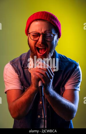 Happy positive hipster man with beard standing with microphone in both hands, singing songs, wearing beanie hat and denim vest. Indoor studio shot isolated on colorful neon light background. Stock Photo