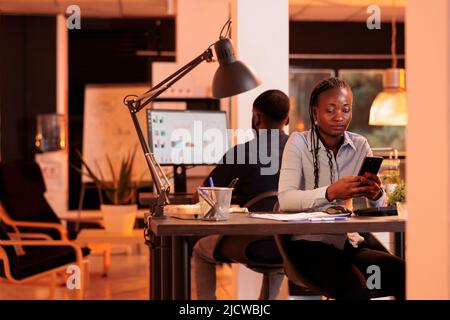 Young businesswoman looking at smartphone app to plan presentation, using social media website to create email report. Analyzing online data on mobile phone to do paperwork research. Stock Photo