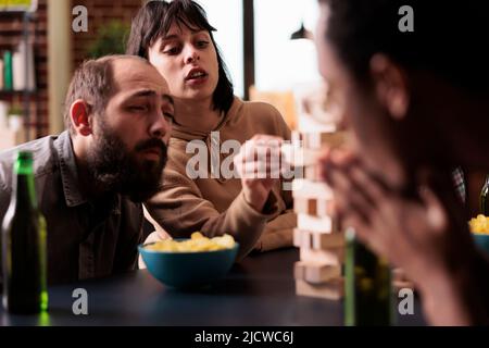 Concentrated woman carefully removing wooden block from wood tower structure. Focused people sitting at table in living room playing society games together while enjoying leisure activity. Stock Photo