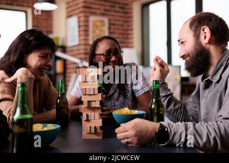 Laughing diverse people sitting at table while talking and enjoying society games together. Multiethnic people sitting at home in living room while discussing and playing with wood blocks. Stock Photo