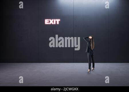 Find right decision and best way-out concept with pensive businesswoman back view in empty room looking at glowing neon exit sign on dark wall Stock Photo